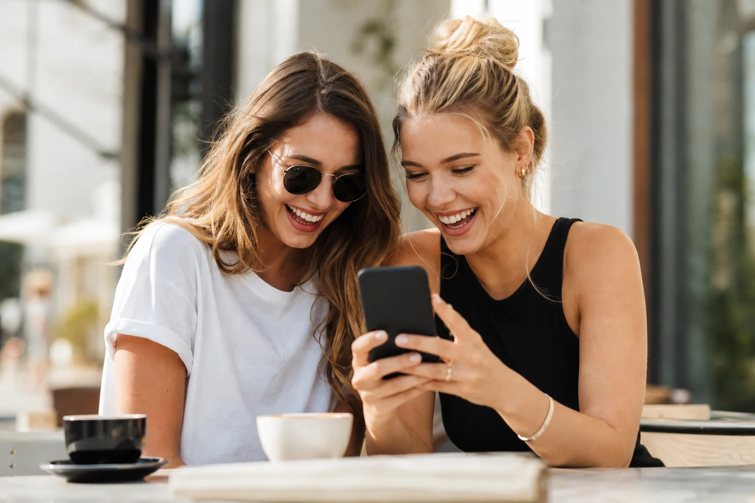 Two friends laughing over a phone at an outdoor cafe, showing a good Tinder match conversation